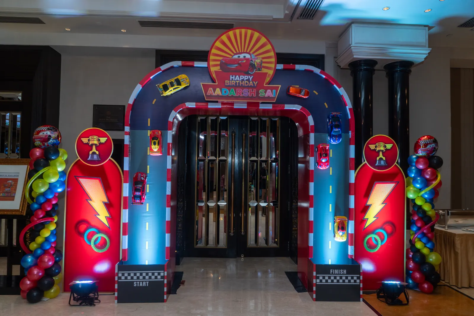 Race-car themed entrance arch with a chequered track, toy cars, start and finish blocks and balloon columns