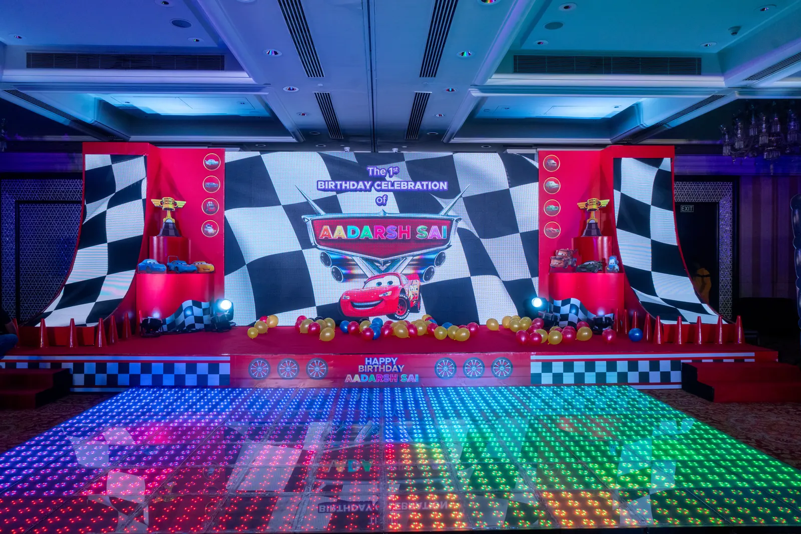 Race-car themed stage with a chequered flag backdrop, pit-stop props, balloon clusters and a full RGB LED dance floor
