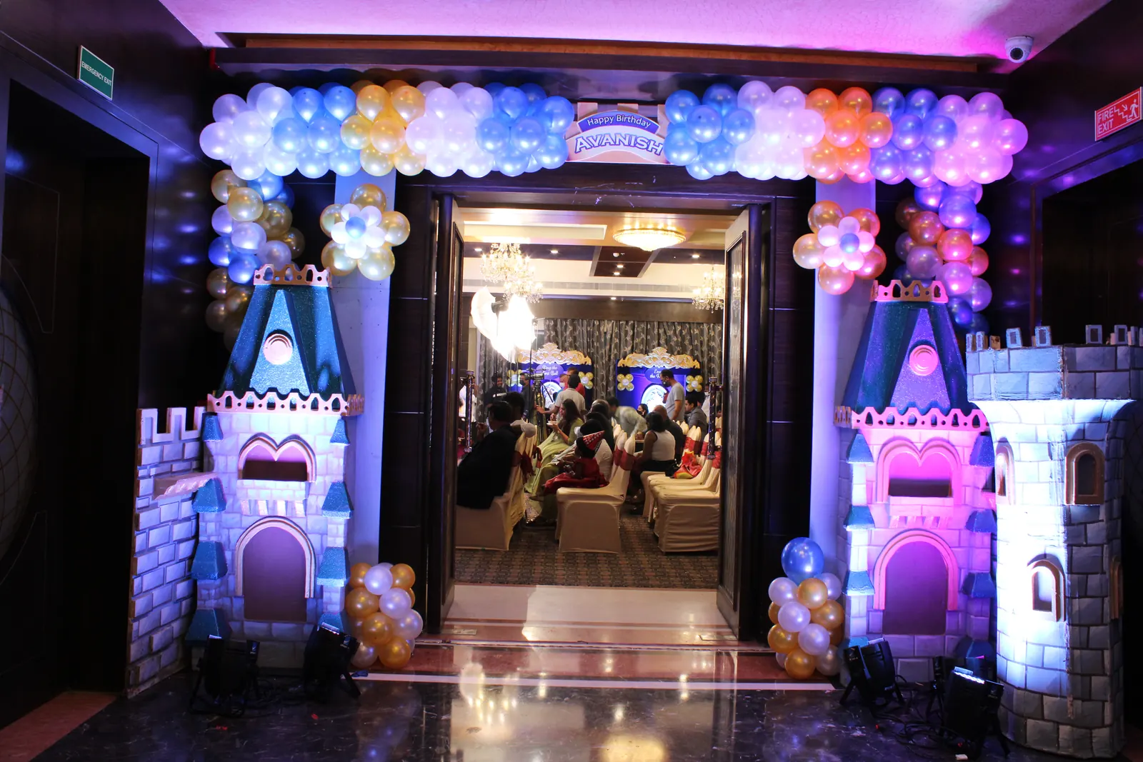 Castle themed entrance with sculpted towers, a pastel balloon garland arch and a lit birthday sign above the doorway