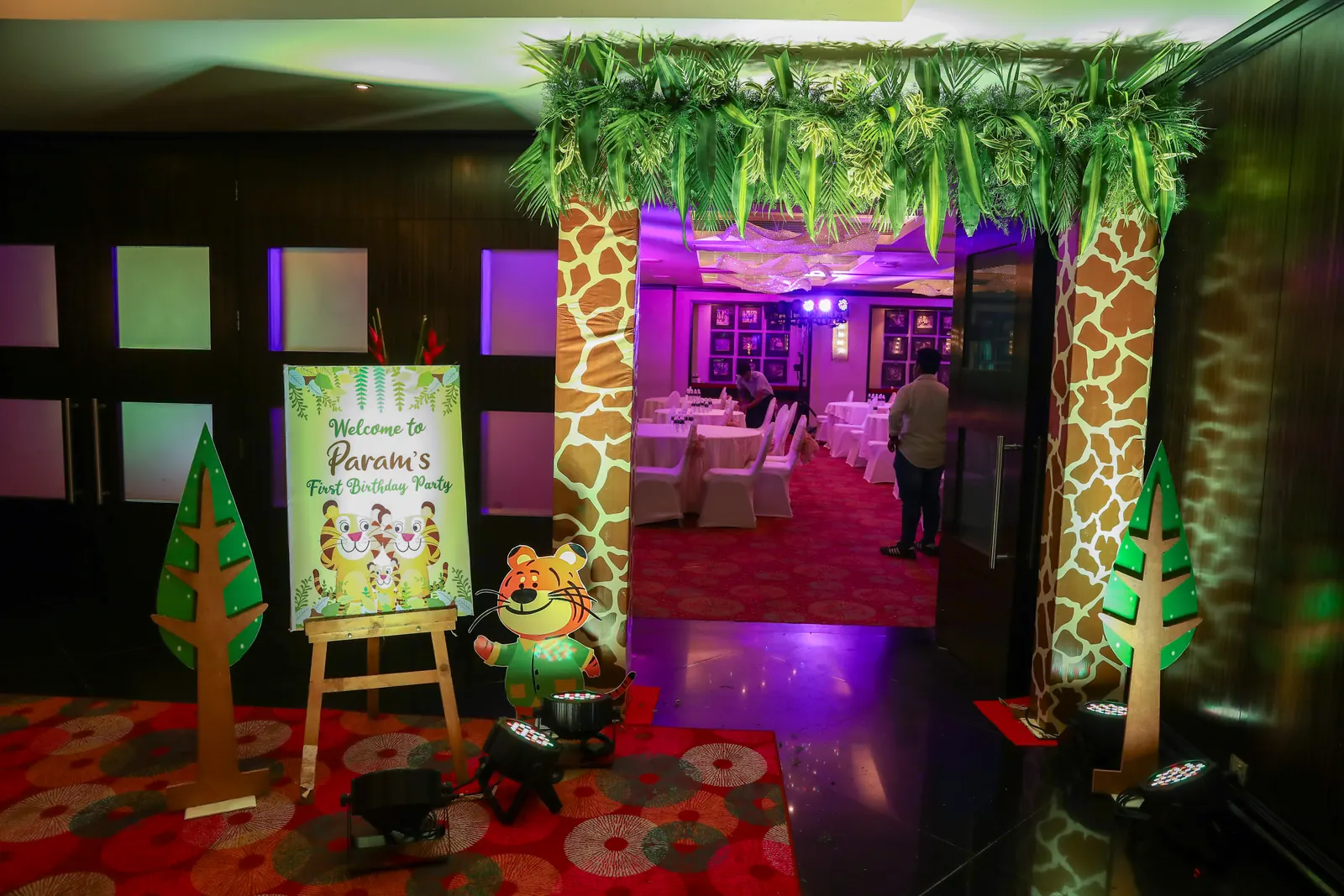 Jungle themed first birthday entrance with giraffe-print pillars, a hanging foliage canopy and a welcome board on an easel