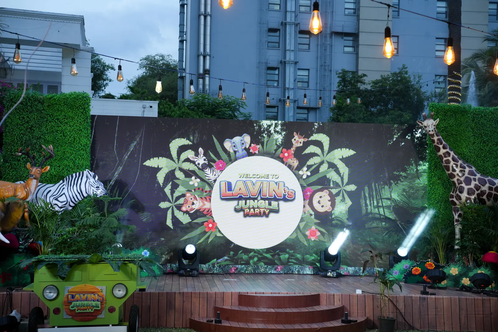 Outdoor jungle themed stage backdrop with a green foliage wall, animal cut-outs and festoon lighting