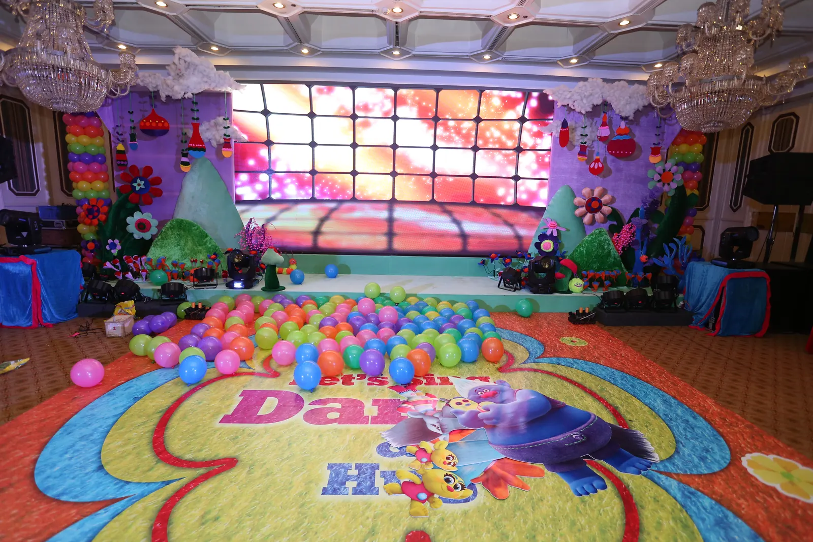 Trolls themed stage with an LED screen, rainbow balloon columns, felt hills and a ball pit spilling onto a printed dance floor
